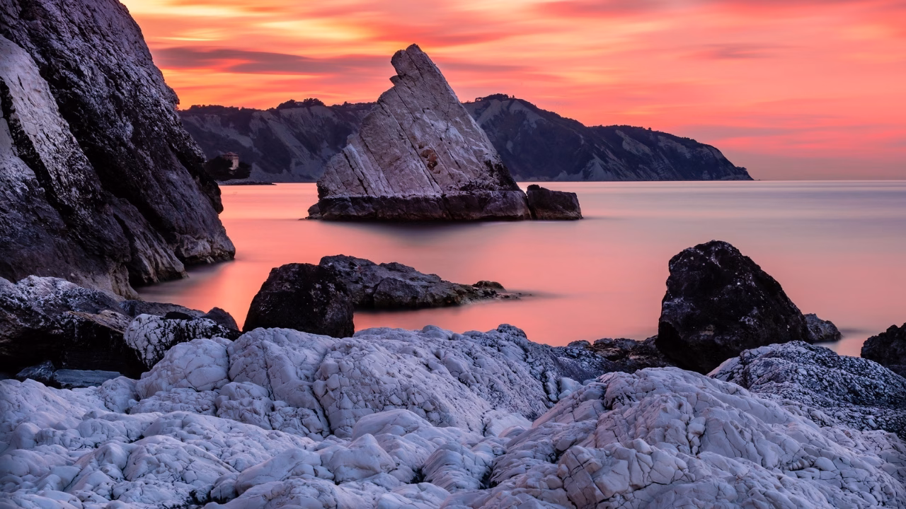 Scogli bianchi frastagliati che emergono dall&rsquo;acqua calma al tramonto, con il caratteristico scoglio &ldquo;La Vela&rdquo; del Conero proprio dalla spiaggia gay La vela del Conero, una delle spiagge LGBTQ delle Marche