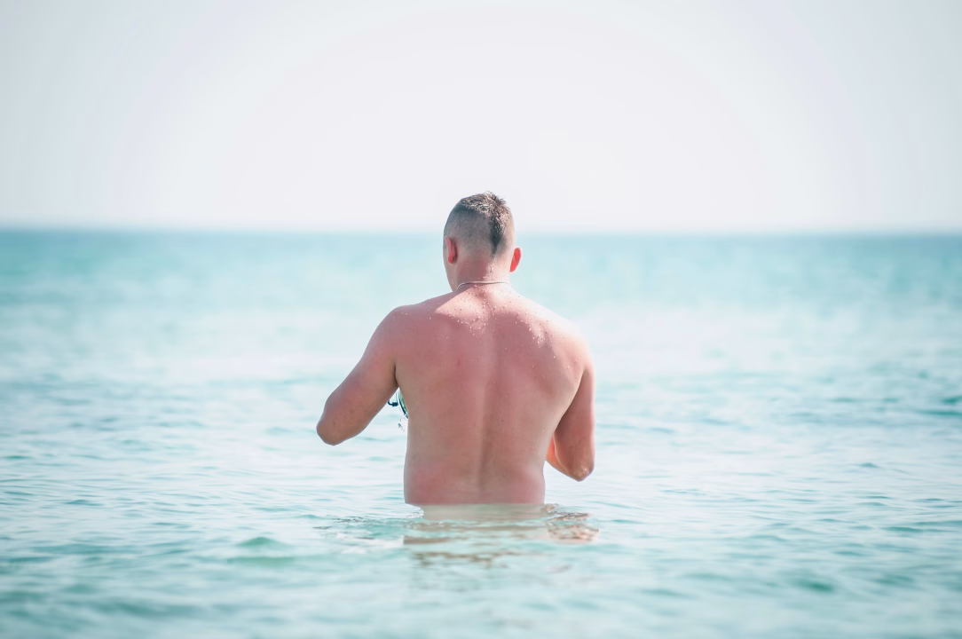 Uomo di spalle immerso nel mare cristallino della spiaggia naturista gay di Pizzogreco, nella riserva marina di Capo Rizzuto in Calabria, una delle destinazioni FKK pi&ugrave; apprezzate dalla comunit&agrave; LGBTQ+ nel sud Italia.