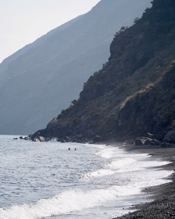 Tratto di costa vulcanica a Stromboli con sabbia nera, onde delicate e montagne scure a picco sul mare; alcune persone si rilassano e nuotano in un angolo appartato della spiaggia.