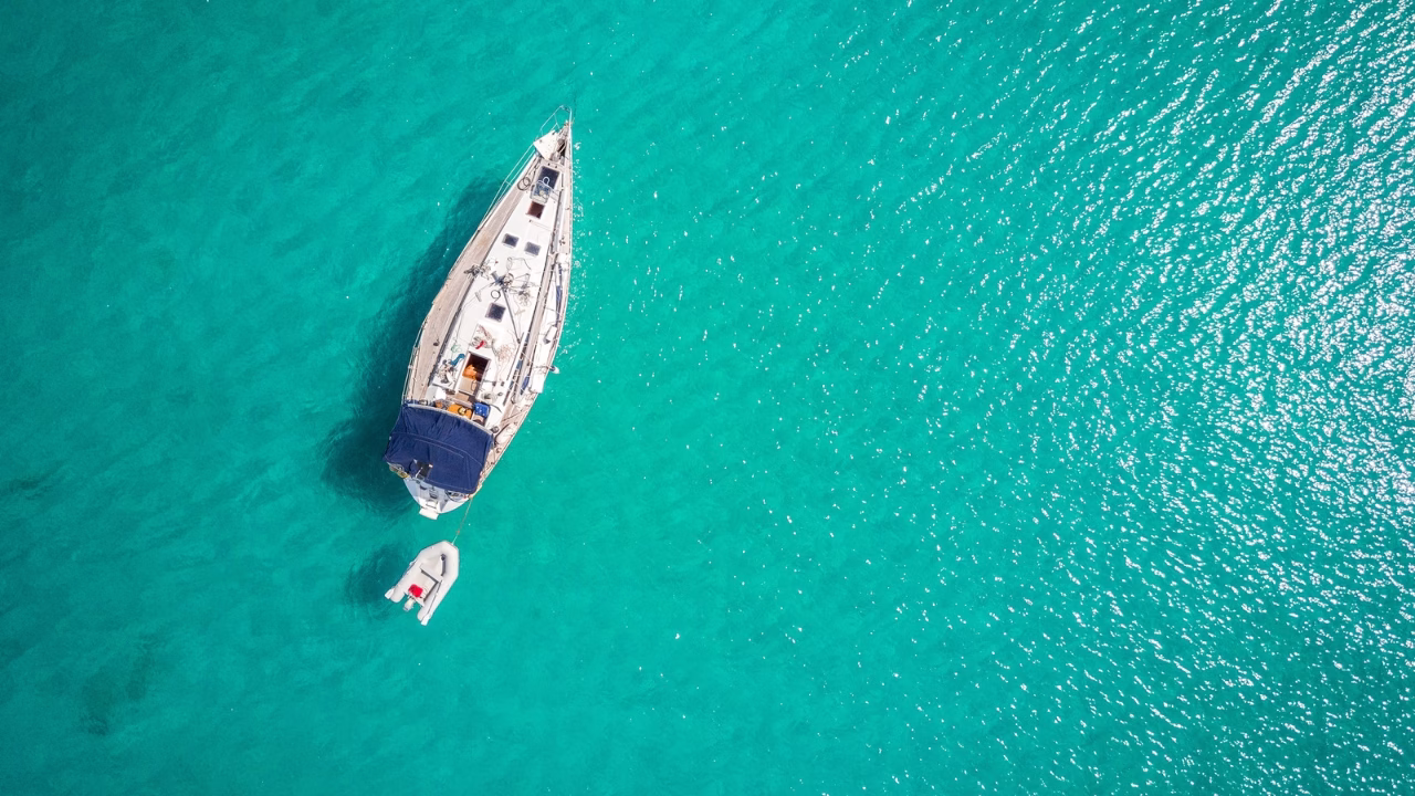 Vista aerea di una barca a vela ancorata nelle acque cristalline della Baia del Mulino d&rsquo;Acqua a Otranto, con un piccolo gommone al seguito. Una destinazione perfetta per un&rsquo;escursione in barca nel Salento, immersi in un mare turchese da sogno.