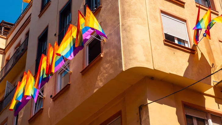Una fila di bandiere arcobaleno sventola da diverse finestre di un edificio color ocra sotto un cielo limpido, simbolo dell&rsquo;accoglienza LGBTQ+ in una struttura probabilmente gay-friendly o naturista a Sitges, una delle destinazioni pi&ugrave; amate dalla comunit&agrave; queer in Spagna.