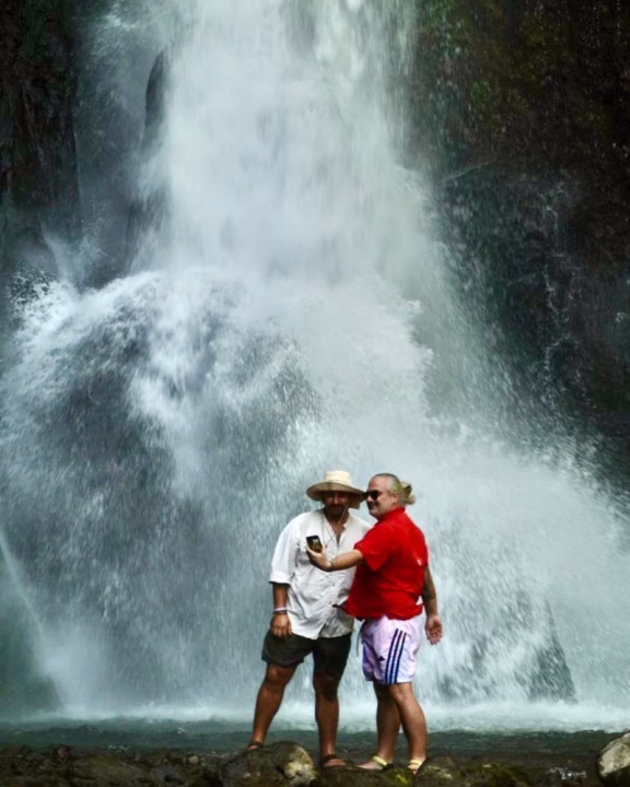 Daniele e Luigi di Gayly Planet in posa davanti alla maestosa cascata di Nungnung a Bali, con l&rsquo;acqua che si infrange rumorosamente sulle rocce. Una delle persone, con una camicia rossa e occhiali da sole, scatta un selfie mentre l&rsquo;altra, con cappello di paglia e camicia bianca, sorride accanto. Il contrasto tra la potenza della natura e il momento di spensieratezza crea un&rsquo;atmosfera suggestiva e avventurosa.