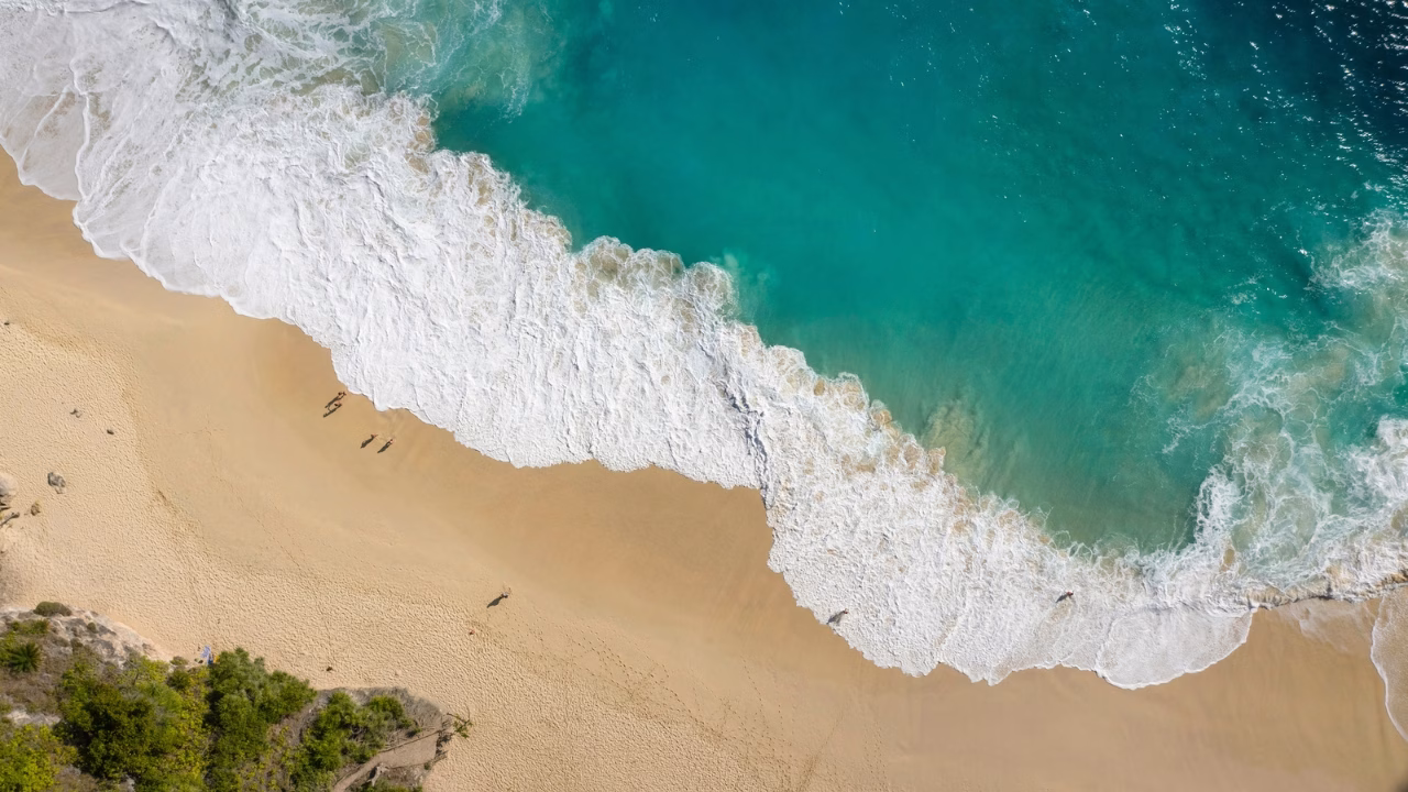 Una veduta aerea mozzafiato di una spiaggia tropicale a Nusa Penida, Bali, con onde turchesi che si infrangono sulla sabbia dorata. Piccole figure di persone camminano lungo la riva, lasciando impronte sulla battigia bagnata. Il contrasto tra il verde della vegetazione, il bianco della schiuma marina e il blu dell’oceano rende l’immagine ancora più suggestiva.