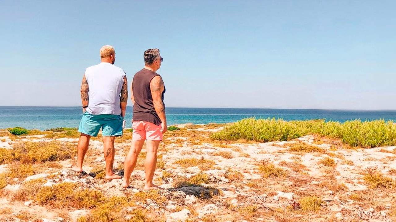 Daniele e Luigi di Gayly Planet in abbigliamento da spiaggia camminano tra le dune della spiaggia gay del Pizzo a Gallipoli, ammirando il mare calmo e limpido sotto un cielo azzurro.