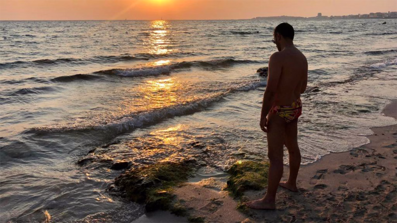 Uomo in costume da bagno osserva il tramonto sul mare dalla spiaggia gay di Gallipoli, con le onde che lambiscono la riva sabbiosa e la luce dorata che si riflette sull&rsquo;acqua. Una delle destinazioni LGBTQ+ pi&ugrave; amate del Salento.