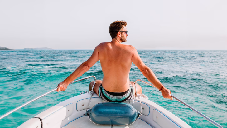 Uomo seduto a poppa su una barca che naviga nel mare cristallino del Salento, con costume a righe e sguardo rivolto verso l&rsquo;orizzonte.