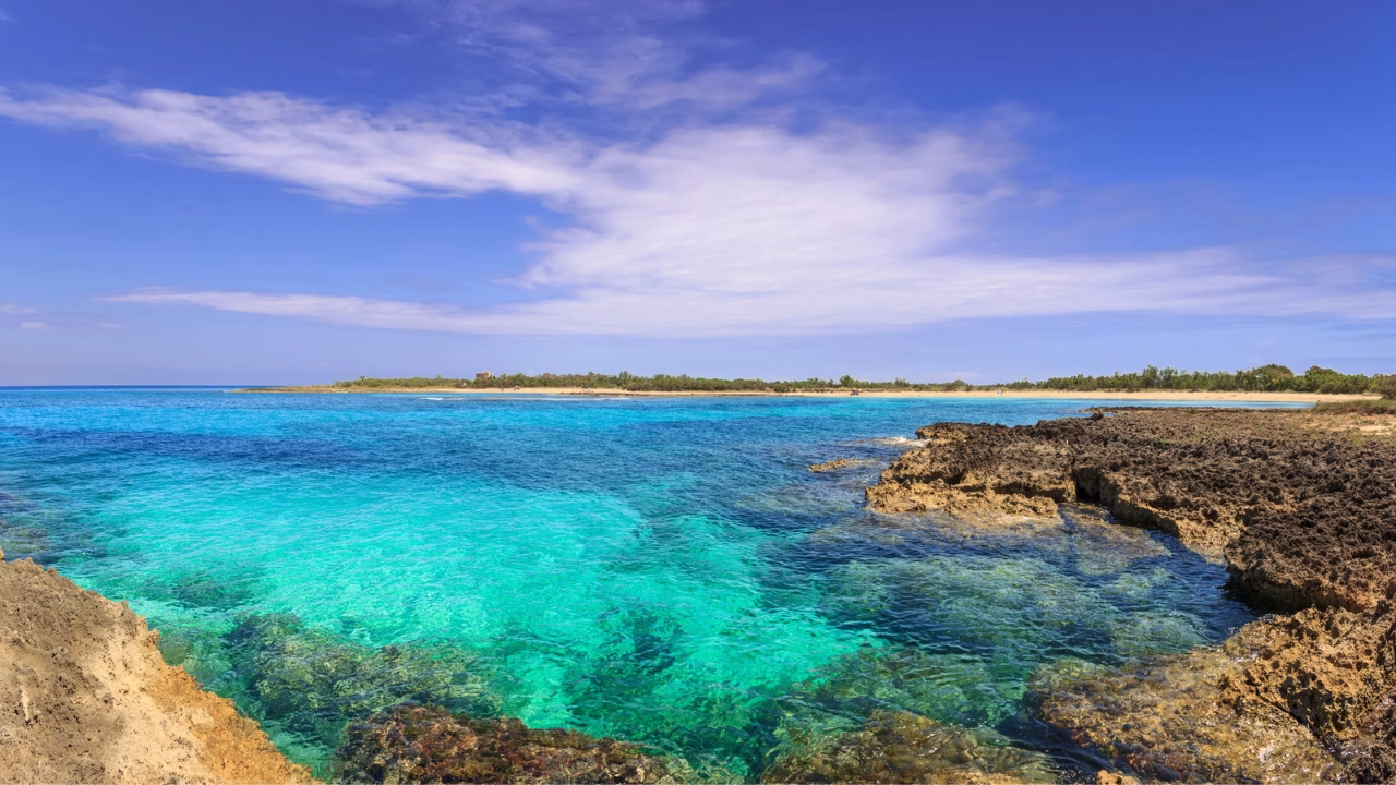 Scorcio panoramico della spiaggia naturista di Torre Guaceto, vicino Brindisi, con acqua cristallina, rocce a picco e vegetazione selvaggia, perfetta per un&rsquo;esperienza gay-friendly in Puglia.