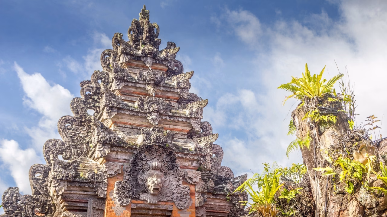 Un primo piano della porta di un tempio balinese a Ubud, Bali, decorata con intricate sculture in pietra che raffigurano motivi tradizionali e figure mitologiche. L’architettura presenta dettagli scolpiti con precisione, tipici dell’arte sacra balinese, con una struttura in pietra rossa e muschio che si fonde con la natura circostante. Sullo sfondo, il cielo azzurro con nuvole bianche e una pianta verde che cresce su un tronco completano la scena.