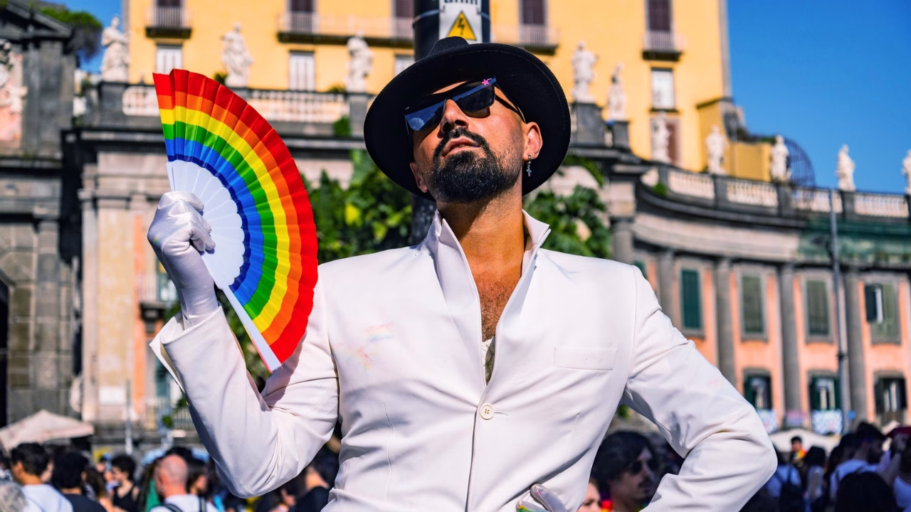 Una persona con barba, occhiali da sole e cappello nero posa con fierezza al Pride di Napoli, indossando un elegante completo bianco e guanti. Tiene in mano un ventaglio arcobaleno, simbolo LGBTQ+, con un’espressione fiera e sicura. Sullo sfondo, edifici storici e una folla festosa.