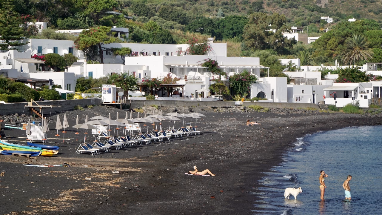La spiaggia di Ficogrande di sabbia nera a Stromboli con lettini e ombrelloni chiusi, affacciata su un mare calmo. In riva, due persone e un cane bianco si godono l&rsquo;acqua, mentre pi&ugrave; indietro una persona prende il sole sulla spiaggia. Sullo sfondo, il pittoresco borgo di Stromboli con edifici bianchi e vegetazione lussureggiante crea un&rsquo;atmosfera mediterranea e rilassante.