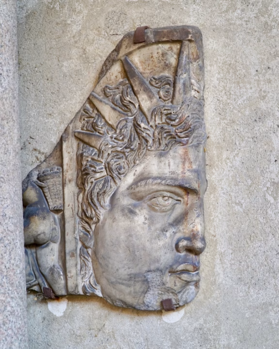 Bassorilievo del Sol Invictus nel Chiostro Cappuccio a Milano, un&rsquo;antica rappresentazione del dio solare romano. La scultura in pietra mostra un volto maschile con una corona raggiata, simbolo di luce e potere divino, incastonata tra le mura storiche del chiostro