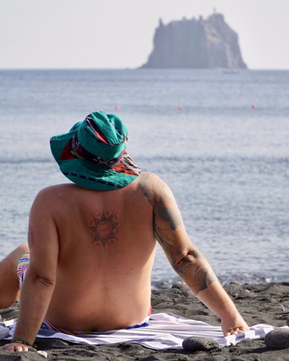 Una persona tatuata, Luigi di Gayly Planet, con cappello verde e costume colorato, &egrave; seduto sulla spiaggia nera di Stromboli guardando il mare e l&rsquo;isolotto di Strombolicchio sullo sfondo. L&rsquo;atmosfera &egrave; rilassata e contemplativa.