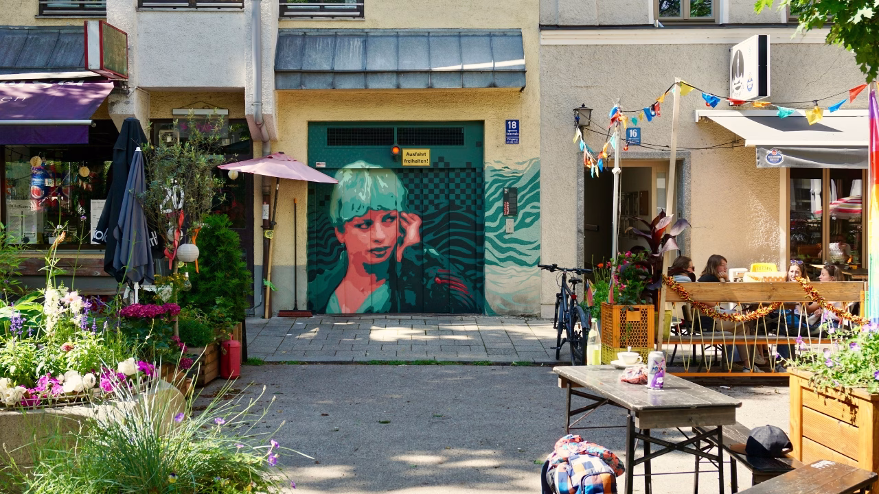 Cortile colorato e vivace nel quartiere gay di Monaco di Baviera, Glockenbachviertel, con murale artistico, fiori e caff&egrave; all&rsquo;aperto, cuore della scena LGBTQ+ della citt&agrave;.