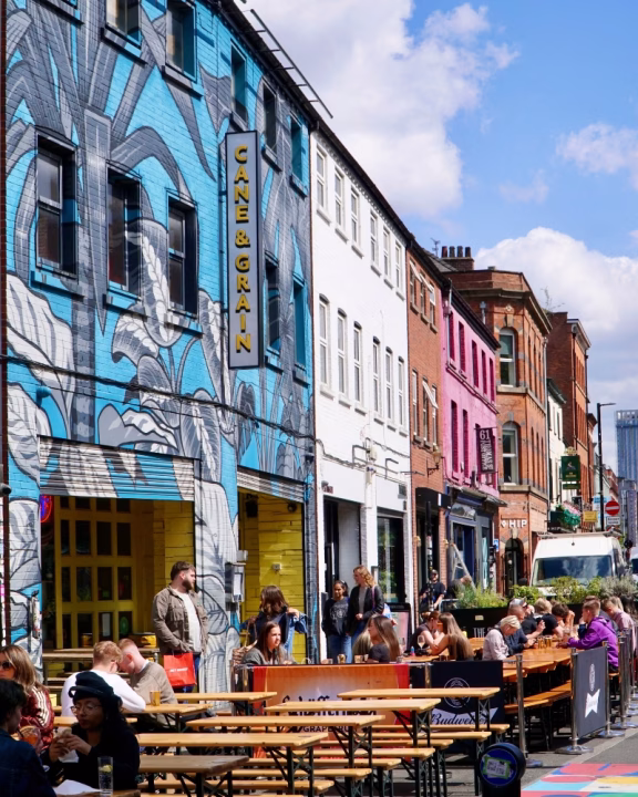 Persone sedute ai tavoli all&rsquo;aperto nel Northern Quarter, quartiere artistico e queer di Manchester, con facciate colorate e murales su edifici storici.