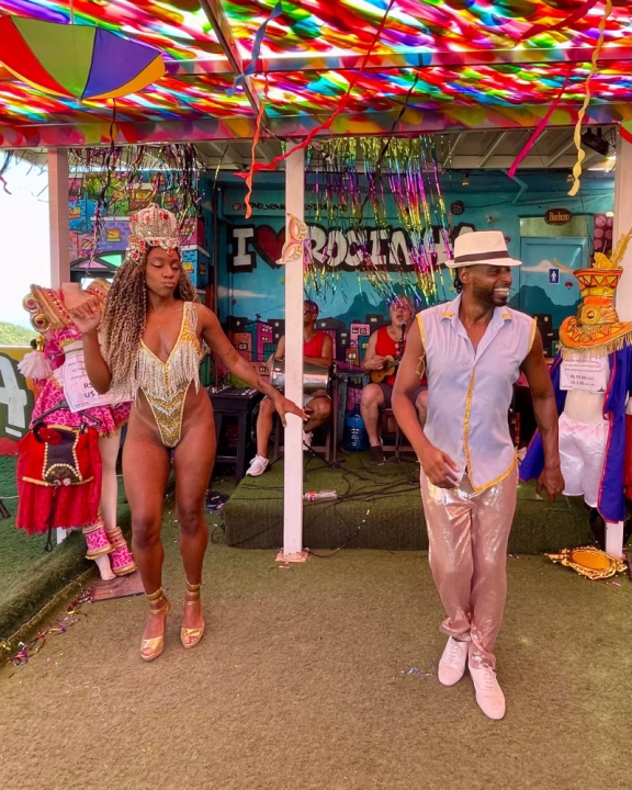 Due ballerini si esibiscono in una coloratissima Roda de Samba queer a Rio de Janeiro, sotto una tettoia decorata con ombrelli arcobaleno, nastri e festoni. La performer indossa un body dorato e una corona, mentre l&rsquo;uomo accanto a lei sorride danzando in pantaloni luccicanti e camicia smanicata. Sullo sfondo si legge &ldquo;I  love Rocinha&rdquo;, indicando la celebre favela.