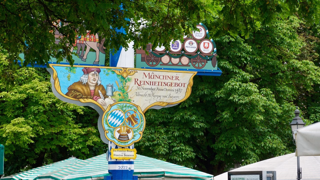 Cartello decorativo nel Biergarten del Viktualienmarkt di Monaco di Baviera che celebra il M&uuml;nchner Reinheitsgebot, l&rsquo;antica legge bavarese sulla purezza della birra emanata nel 1487, circondato da alberi e tende del mercato.