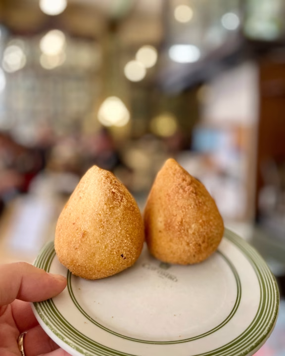 Due coxinha dorate, una tipica specialit&agrave; brasiliana a forma di goccia, servite su un piattino da bar con bordo verde. L&rsquo;immagine &egrave; scattata in un locale tradizionale di Rio de Janeiro, sfocato sullo sfondo, probabilmente durante un&rsquo;esperienza gastronomica queer-friendly.