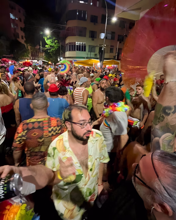Una folla vivace si raduna in strada durante una festa notturna LGBTQ+ a Rio de Janeiro, con persone che ballano, indossano abiti colorati e accessori arcobaleno. L&rsquo;atmosfera &egrave; elettrica e inclusiva, tra musica, drink e celebrazione queer sotto le luci della citt&agrave;.