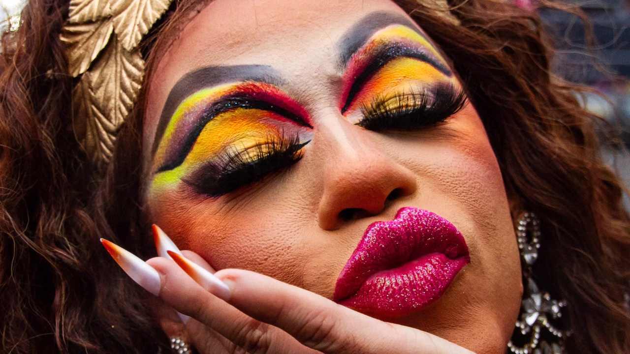 Primo piano mozzafiato di una drag queen con trucco scenografico: occhi sfumati nei toni del giallo, rosso e nero, ciglia voluminose e labbra glitterate fucsia. L&rsquo;espressione &egrave; intensa e teatrale, incarnando lo spirito vibrante dei drag show LGBTQ+ a Rio de Janeiro.