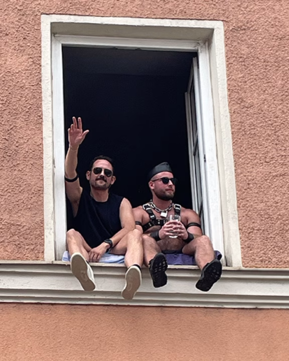 Due uomini seduti su un davanzale aperto a Monaco di Baviera durante un evento LGBTQ+, uno con abbigliamento casual e l&rsquo;altro con harness in stile fetish leather, simbolo della scena queer alternativa e dei club fetish della citt&agrave;.