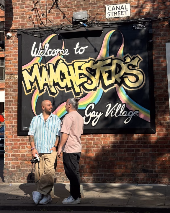 Daniele e Luigi di Gayly Planet si incontrano davanti al murale &ldquo;Welcome to Manchester&rsquo;s Gay Village&rdquo; su Canal Street, cuore della vita LGBTQ+ a Manchester, con scritta arcobaleno e atmosfera inclusiva nel celebre quartiere gay della citt&agrave;.