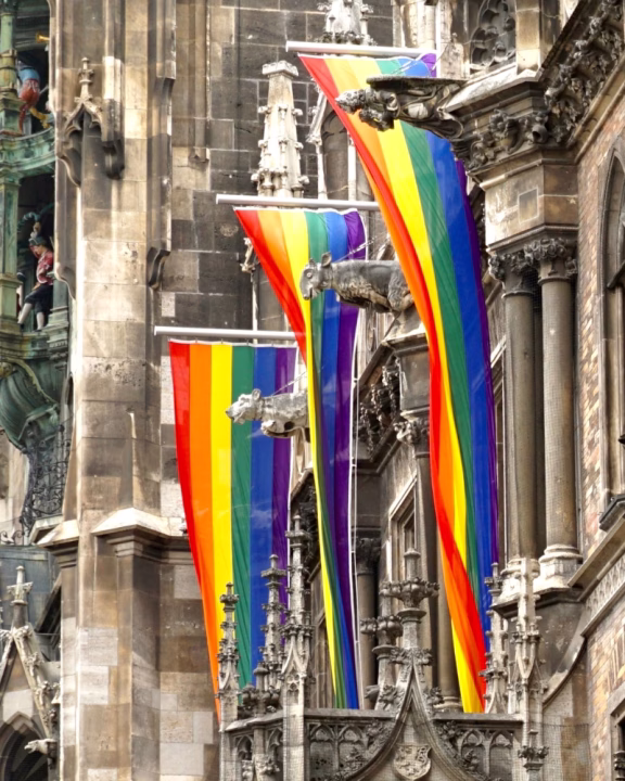 Bandiere arcobaleno sventolano dalle guglie gotiche del municipio di Monaco di Baviera durante il Pride, drappeggiate tra gargoyle e dettagli in pietra scolpita, simbolo del sostegno della citt&agrave; alla comunit&agrave; LGBTQ+.