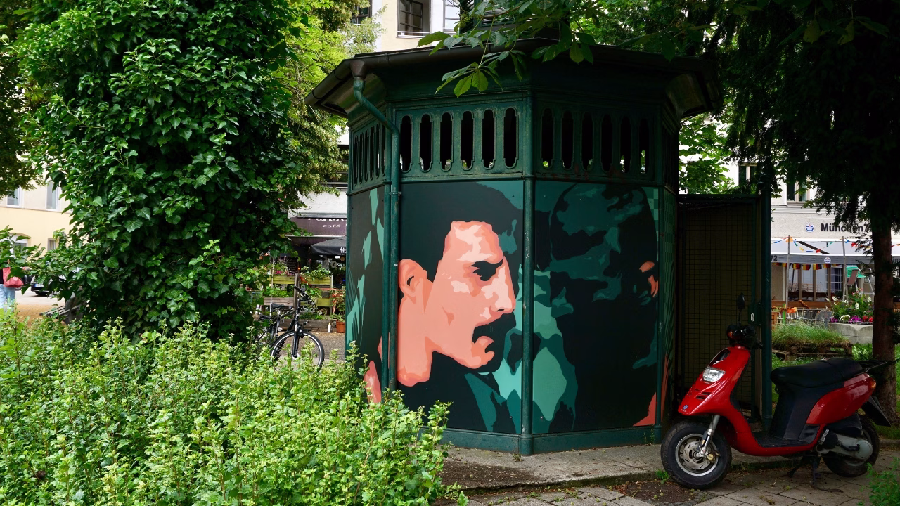 Murale di Freddie Mercury dipinto su una vecchia struttura urbana nel verde Holzplatz di Monaco di Baviera, omaggio iconico alla cultura LGBTQ+ in uno dei quartieri pi&ugrave; queer-friendly della citt&agrave;.