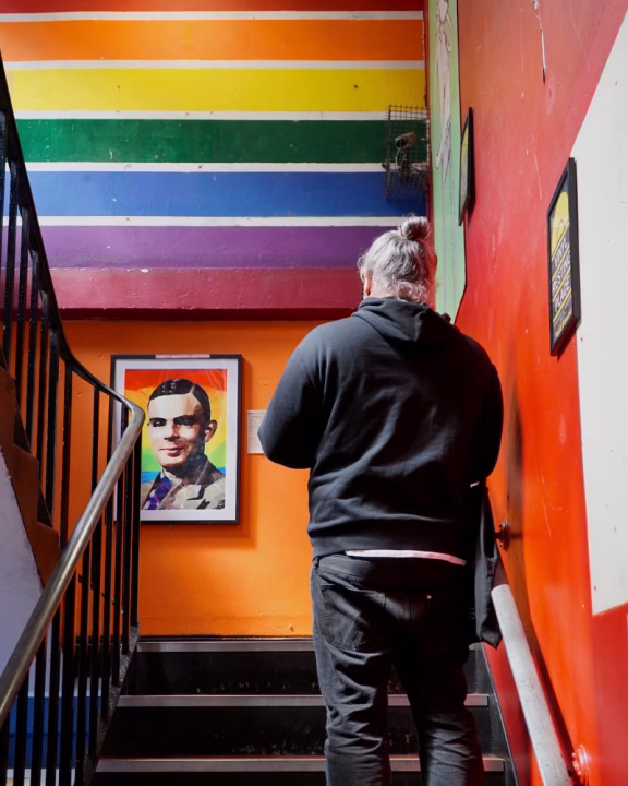 Interno colorato del negozio queer Afflecks a Manchester con bandiera arcobaleno dipinta sul muro e ritratto di Alan Turing, icona LGBTQ+, osservato da un visitatore in cima alle scale.