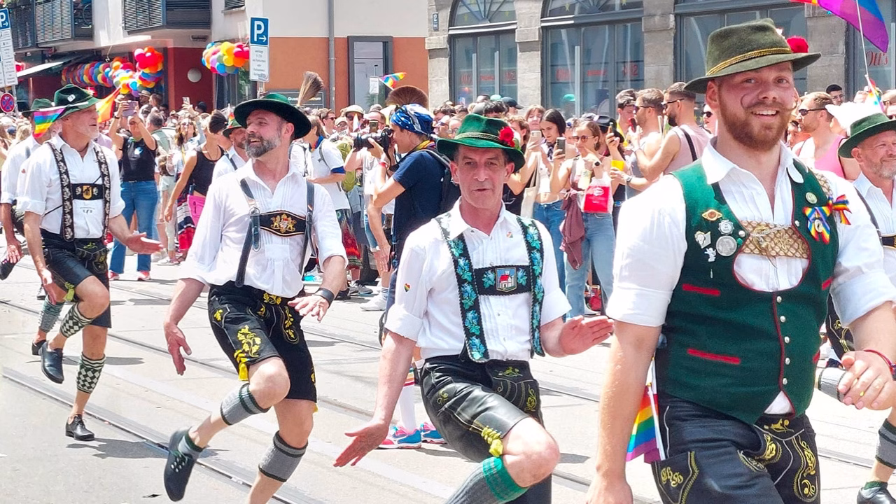 Gruppo di uomini in abiti tradizionali bavaresi sfila durante la Rosa Wiesn, l&rsquo;Oktoberfest gay di Monaco di Baviera, celebrando l&rsquo;orgoglio LGBTQ+ con bandiere arcobaleno, sorrisi e danze folkloristiche in un contesto festoso e inclusivo.