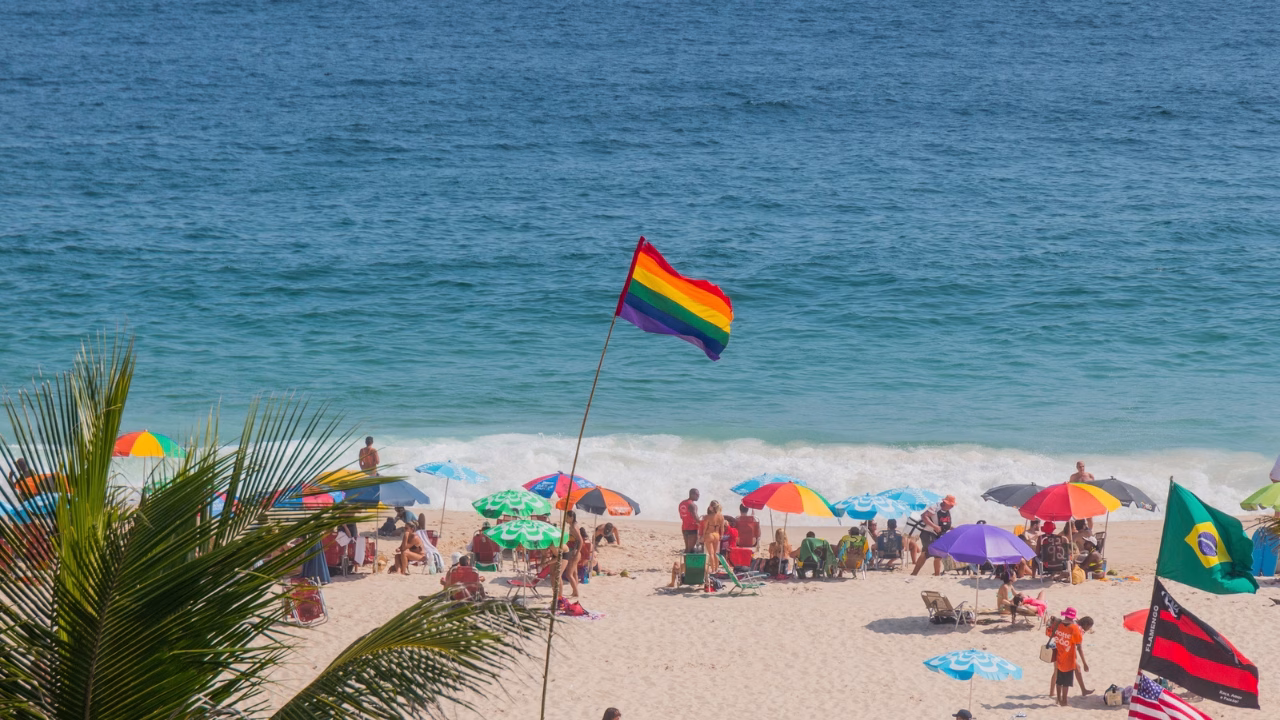 Una colorata spiaggia a Copacabana, Rio de Janeiro, con numerosi ombrelloni variopinti e una bandiera arcobaleno che sventola in primo piano, simbolo dell&rsquo;inclusivit&agrave; LGBTQ+. L&rsquo;oceano azzurro sullo sfondo e le palme inquadrate lateralmente completano la scena vivace e accogliente.