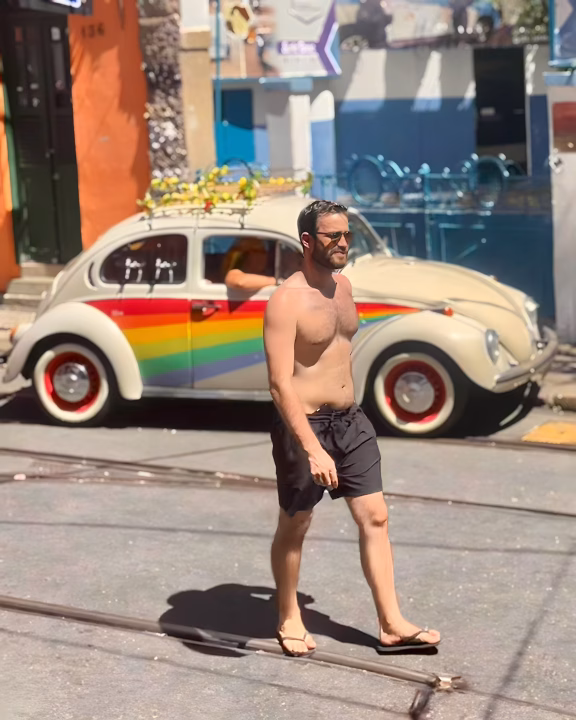 Un uomo a torso nudo cammina con disinvoltura lungo una strada assolata nel quartiere di Santa Teresa a Rio de Janeiro, mentre sullo sfondo si nota un Maggiolino vintage decorato con una vistosa bandiera arcobaleno. L&rsquo;atmosfera rilassata e colorata riflette il carattere boh&eacute;mien e inclusivo di questa zona gay-friendly della citt&agrave;.