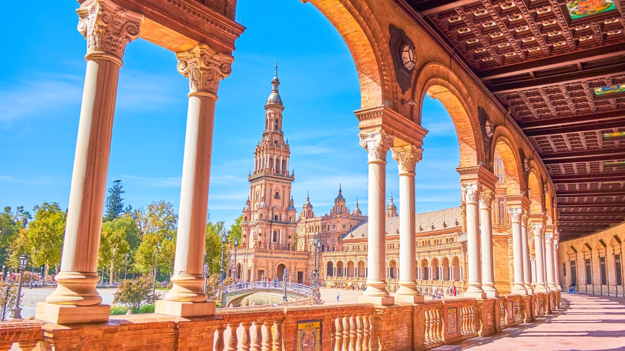 Veduta soleggiata della Plaza de Espa&ntilde;a a Siviglia, Spagna, con le sue maestose arcate in stile rinascimentale e la torre centrale che si erge sullo sfondo. La scena evoca un&rsquo;atmosfera vivace e accogliente, perfetta per un itinerario LGBTQ+ tra arte, cultura e inclusivit&agrave; nel sud della Spagna.