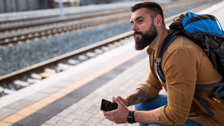 Un uomo con barba e zaino da trekking siede su una banchina ferroviaria, guardando pensieroso verso i binari mentre tiene in mano lo smartphone. L&rsquo;immagine trasmette un senso di attesa e libert&agrave;, perfetta per rappresentare viaggi LGBTQ+ in treno in Spagna.