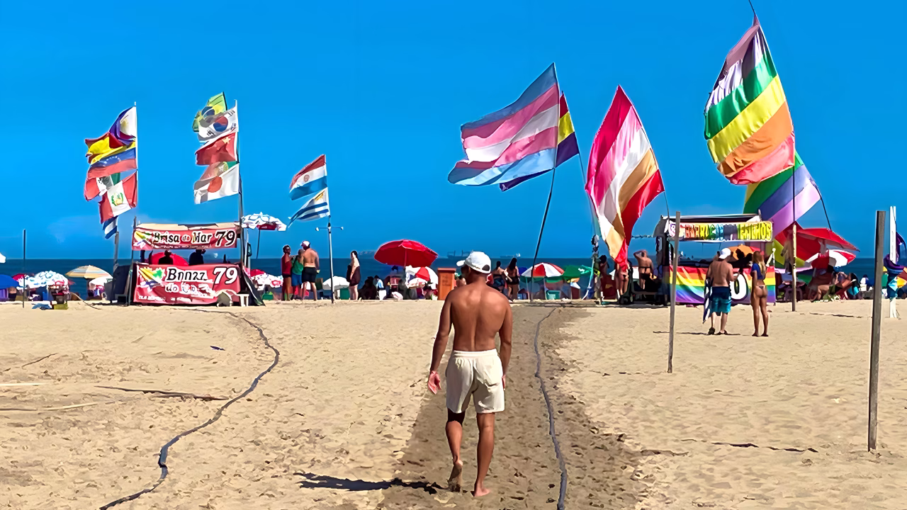 Un uomo cammina verso il mare lungo la spiaggia gay di Copacabana, fermandosi all&rsquo;altezza del punto 79 decorato con grandi bandiere arcobaleno, trans e progress. Una scena colorata e festosa che celebra l&rsquo;inclusivit&agrave; e la cultura LGBTQ+ a Rio de Janeiro.
