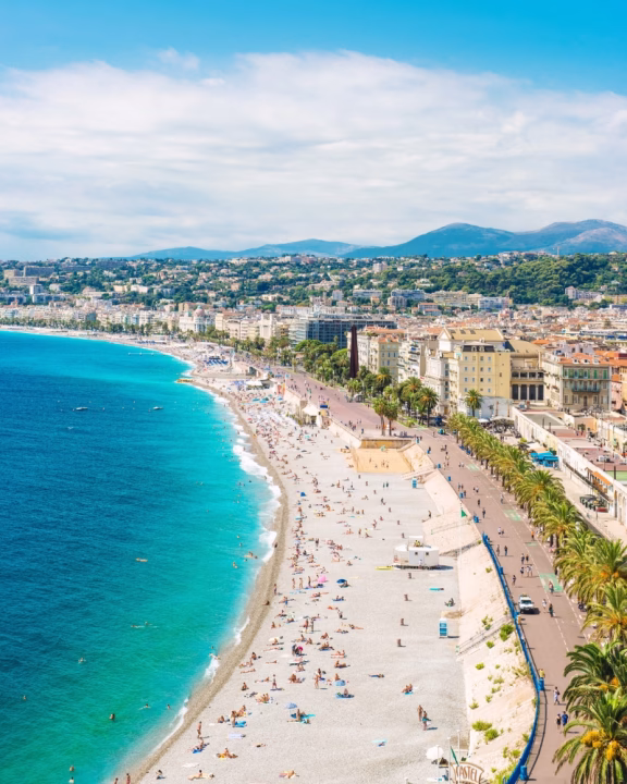 Veduta panoramica della Promenade des Anglais e della spiaggia di Castel Plage a Nizza, con acqua turchese e bagnanti distesi sulla spiaggia ghiaiosa. Lungo la costa si susseguono edifici storici, palme e una passeggiata vivace, ideale per chi cerca spiagge gay-friendly nel cuore della Costa Azzurra.