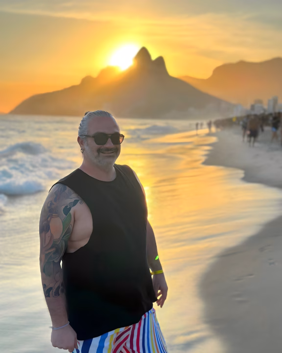 Luigi di Gayly Planet sorridente posa sulla riva della spiaggia di Ipanema al tramonto, con il celebre Morro Dois Irm&atilde;os sullo sfondo e la luce dorata che si riflette sull&rsquo;oceano. Indossa occhiali da sole e pantaloncini arcobaleno, celebrando lo spirito LGBTQ+ in una delle spiagge pi&ugrave; iconiche e inclusive di Rio de Janeiro.