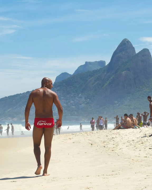 Un uomo cammina lungo la sabbia chiara della spiaggia di Ipanema a Rio de Janeiro, indossando un costume rosso con la scritta &ldquo;Carioca&rdquo;, mentre sullo sfondo spiccano le iconiche montagne Dois Irm&atilde;os e una folla rilassata di bagnanti. L&rsquo;atmosfera &egrave; soleggiata e vibrante, emblematica del carattere inclusivo e vivace della zona LGBTQ+ di Ipanema.