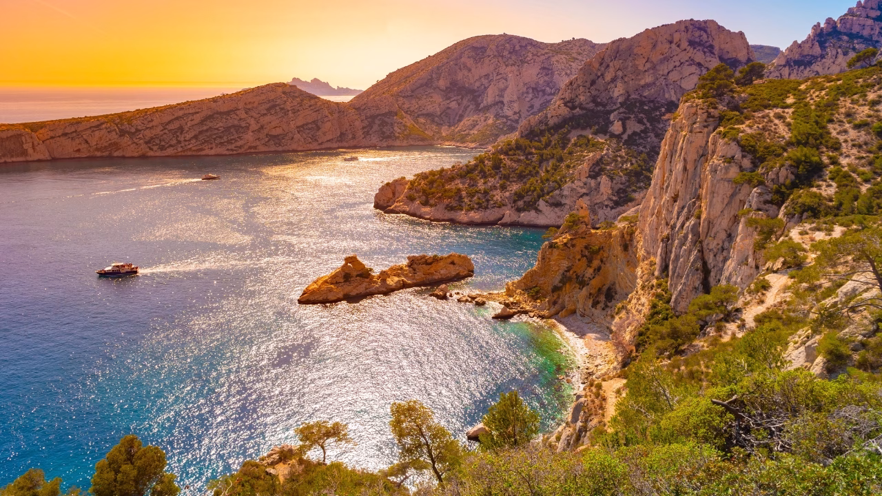 Vista panoramica della Calanque de Sugiton al tramonto, con scogliere rocciose, barche in mare e l&rsquo;acqua che riflette i toni dorati del sole calante. La cala appartata &egrave; una delle spiagge gay e nudiste pi&ugrave; scenografiche del sud della Francia, vicino a Marsiglia.