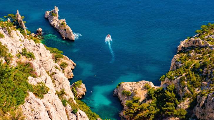 Veduta aerea della Calanque de Sugiton vicino a Marsiglia, con scogliere rocciose bianche ricoperte di vegetazione e mare turchese intenso, dove una piccola barca lascia una scia nell&rsquo;acqua limpida. Una delle spiagge gay e naturiste pi&ugrave; suggestive del sud della Francia.
