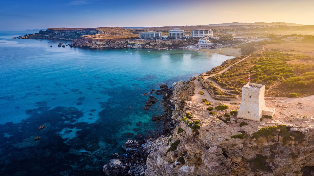 Veduta aerea di Għajn Tuffieħa (Riviera Bay) a Malta con una costa rocciosa con un mare turchese e cristallino, dove si distingue una torre di avvistamento in pietra su un promontorio. In lontananza, spiagge sabbiose e moderni complessi alberghieri si affacciano sulla baia, illuminata da una calda luce al tramonto.