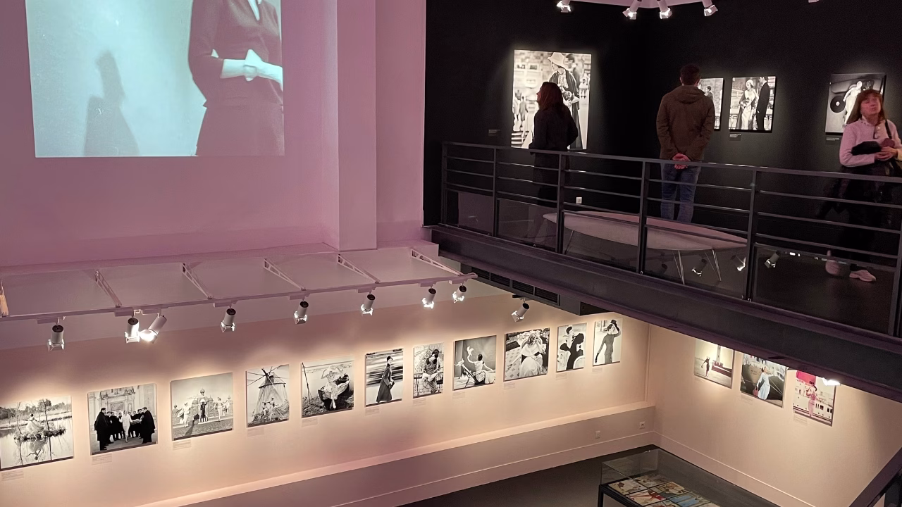 Interno del Mus&eacute;e de la Photographie Charles N&egrave;gre a Nizza, con pareti adornate da fotografie in bianco e nero, proiezione murale di una donna e visitatori che osservano le opere lungo una passerella sopraelevata. Atmosfera intima e artistica in uno spazio espositivo moderno.