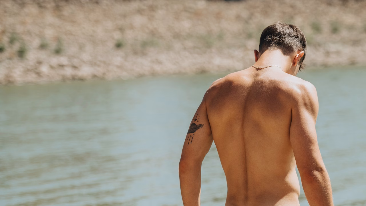 Ragazzo di spalle completamente nudo in riva al mare, con un tatuaggio nero stilizzato sul braccio e la testa leggermente chinata. L&rsquo;immagine rappresenta l&rsquo;atmosfera rilassata e naturale della Plage du Mont Rose, una spiaggia gay e nudista a Marsiglia.