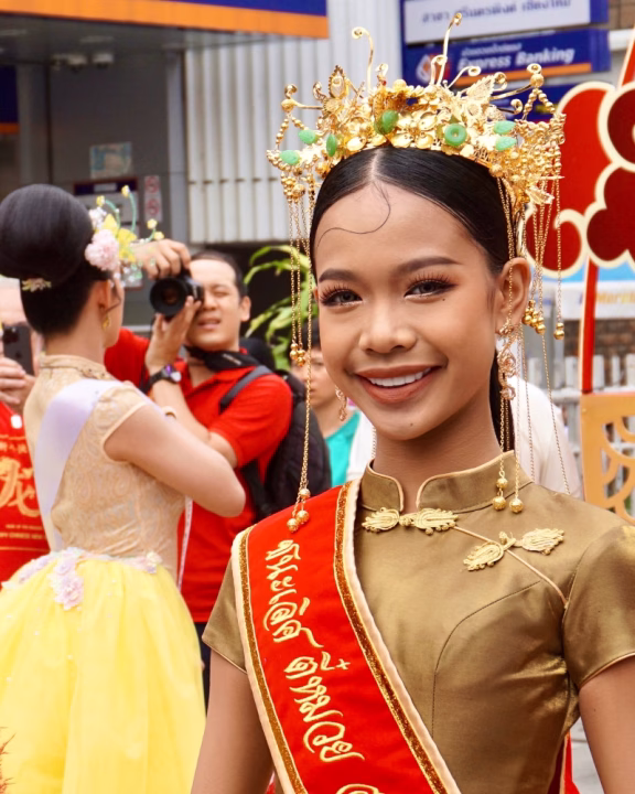 Giovane donna sorridente in abito tradizionale dorato con una fascia rossa decorata. Indossa un elaborato copricapo dorato con dettagli verdi e dorati, durante la festa dei fiori a Chiang Mai in Thailandia.