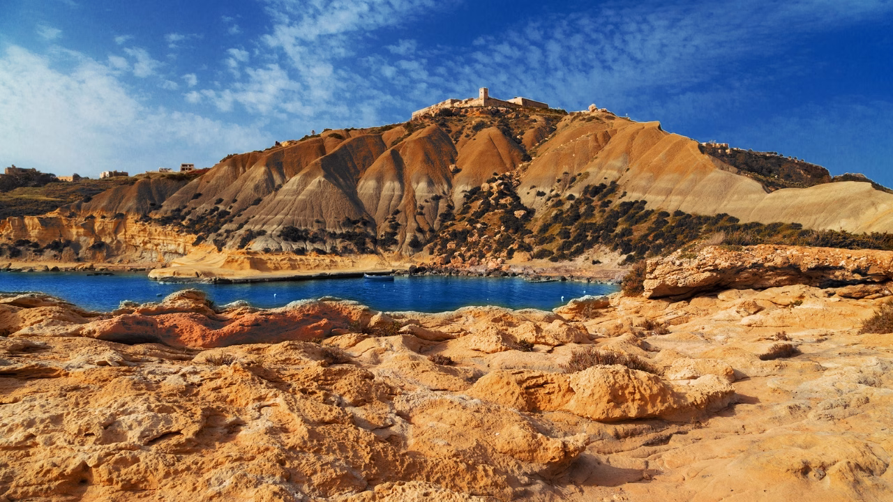 Scogliera rocciosa color ocra sull'isola di Gozo a Malta. La scogliera si affaccia su un&rsquo;insenatura di mare blu intenso, con colline aride e ondulate sullo sfondo. In cima alla collina si intravedono edifici storici e una torre sotto un cielo azzurro con leggere nuvole bianche.