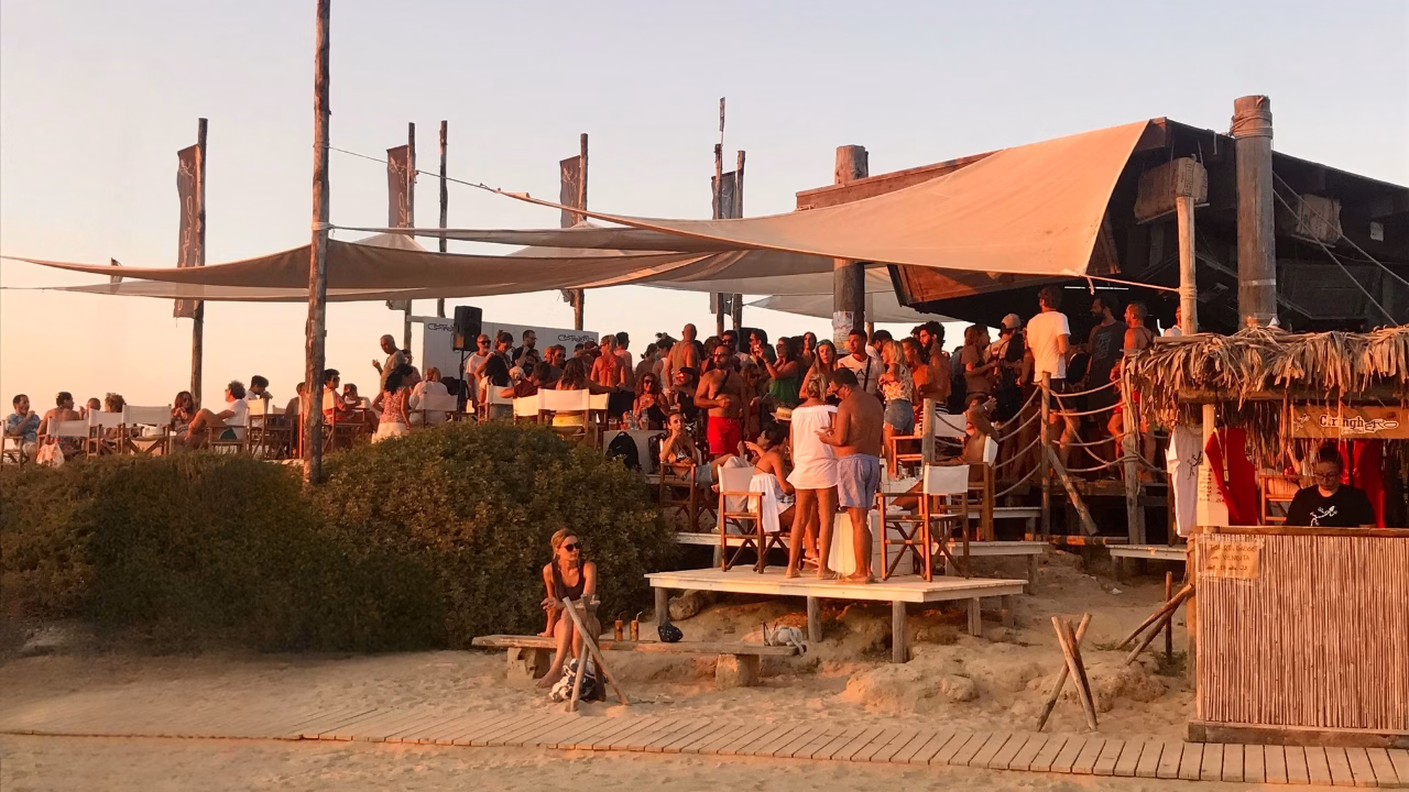 Chiringuito in legno affacciato sulla spiaggia a Punta Pizzo, con una folla di persone che si gode l&rsquo;aperitivo al tramonto sotto tendoni bianchi. L&rsquo;atmosfera &egrave; vivace e informale, con il cielo dorato tipico del Salento che colora l&rsquo;intera scena.