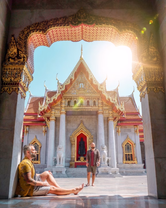 Daniele e Luigi di Gayly Planet in posa davanti al Wat Benchamabophit, il tempio di marmo di Bangkok, con decorazioni dorate e tetto rosso brillante, illuminato dal sole splendente nel cielo azzurro.