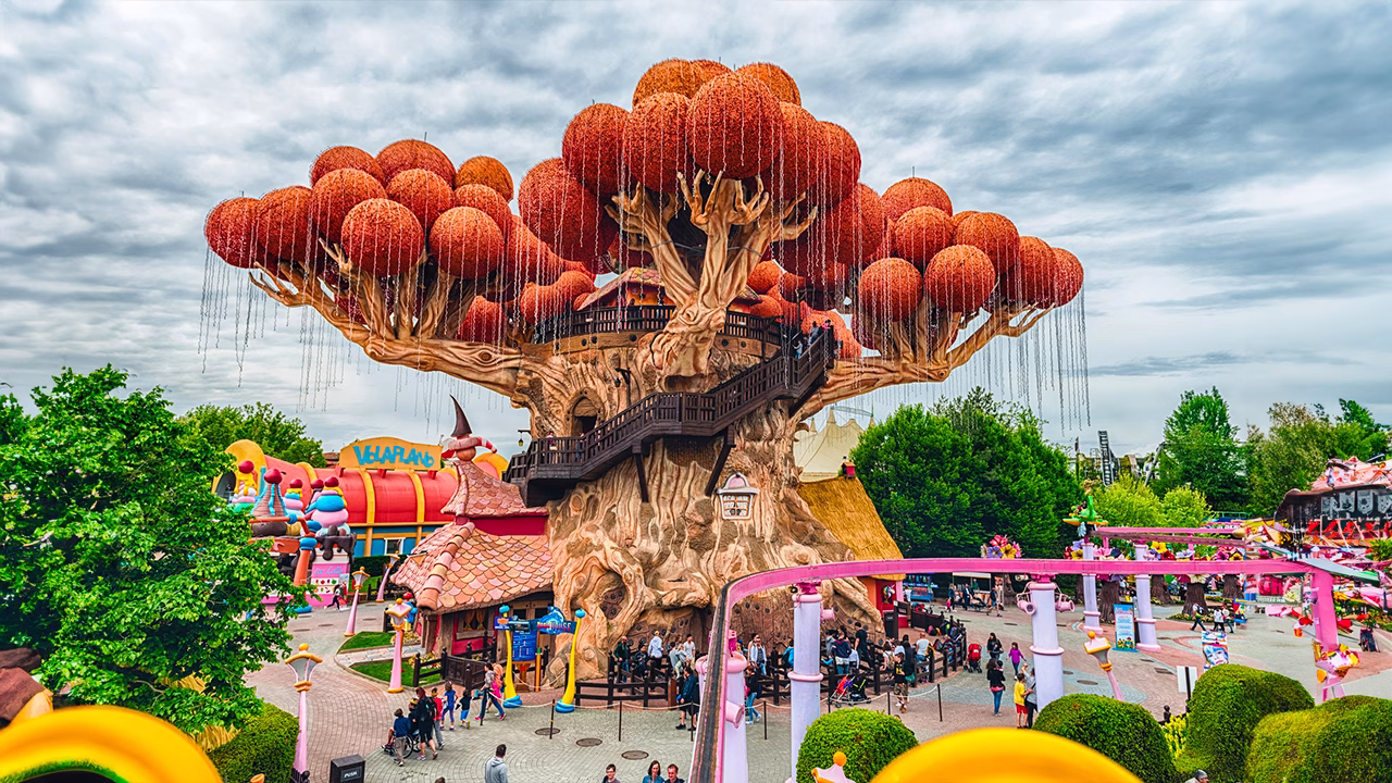 Vista dell&rsquo;albero simbolo di Gardaland, con rami imponenti e sfere arancioni decorative, circondato da visitatori che passeggiano nel parco divertimenti pi&ugrave; famoso d&rsquo;Italia.