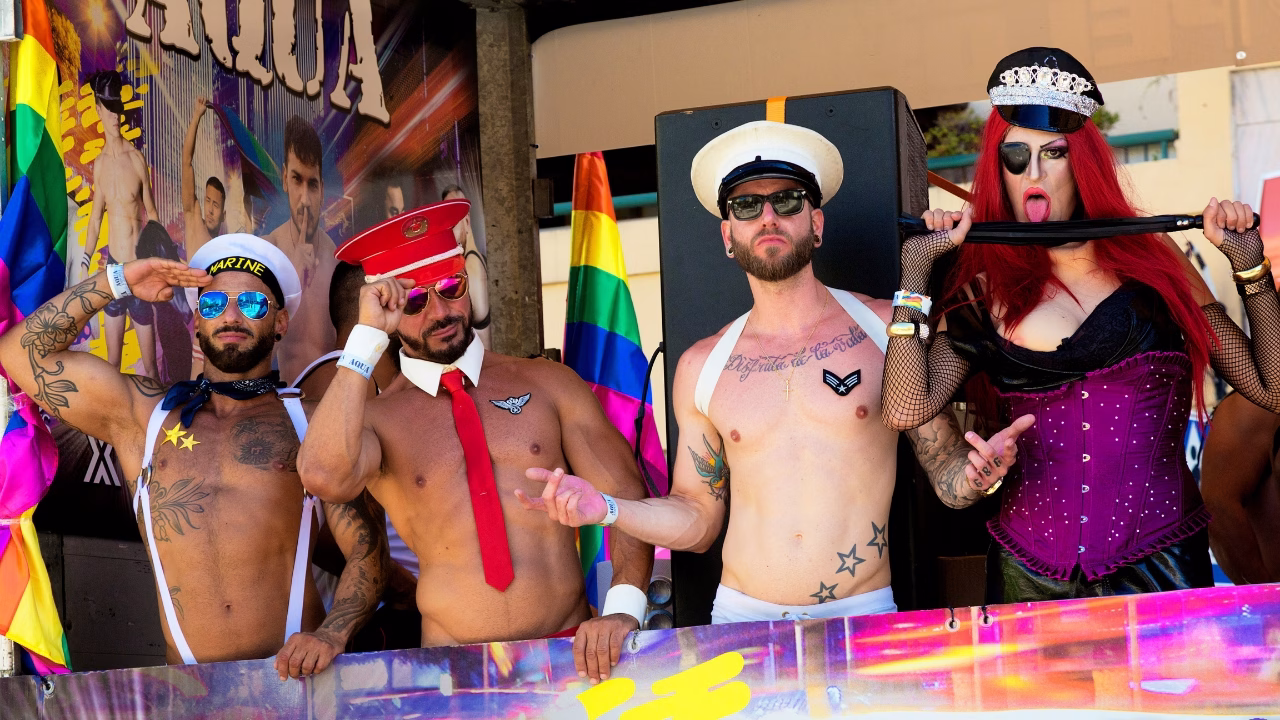 Quattro persone su un carro del Torremolios Pride, tre uomini a torso nudo con costumi da ufficiali sexy e una drag queen con parrucca rossa, bustier viola e cappello da poliziotta. Sullo sfondo, bandiere arcobaleno e un poster colorato animano l’atmosfera festosa.