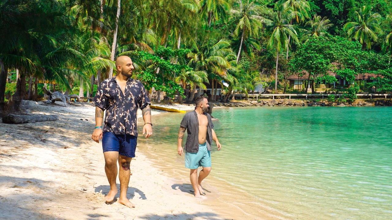 Daniele e Luigi di Gayly Planet camminano a piedi nudi lungo una spiaggia tropicale di Koh Chang in Thailandia, con acqua turchese e palme rigogliose sullo sfondo. Indossano camicie leggere e pantaloncini, godendosi una giornata soleggiata in un ambiente paradisiaco.