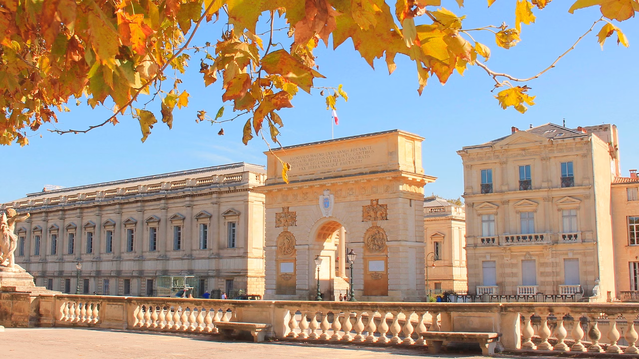 Veduta autunnale dell&rsquo;Arco di Trionfo di Montpellier in Francia con foglie dorate in primo piano e cielo sereno, perfetta meta per un viaggio gay in autunno.